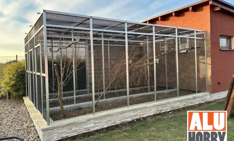 Velká venkovní voliéra pro papoušky s větvemi u domu.
