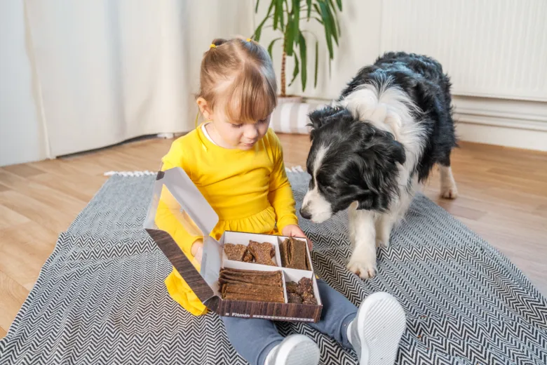 Dívka s psem sedí na koberci u krabice psích sušenek.